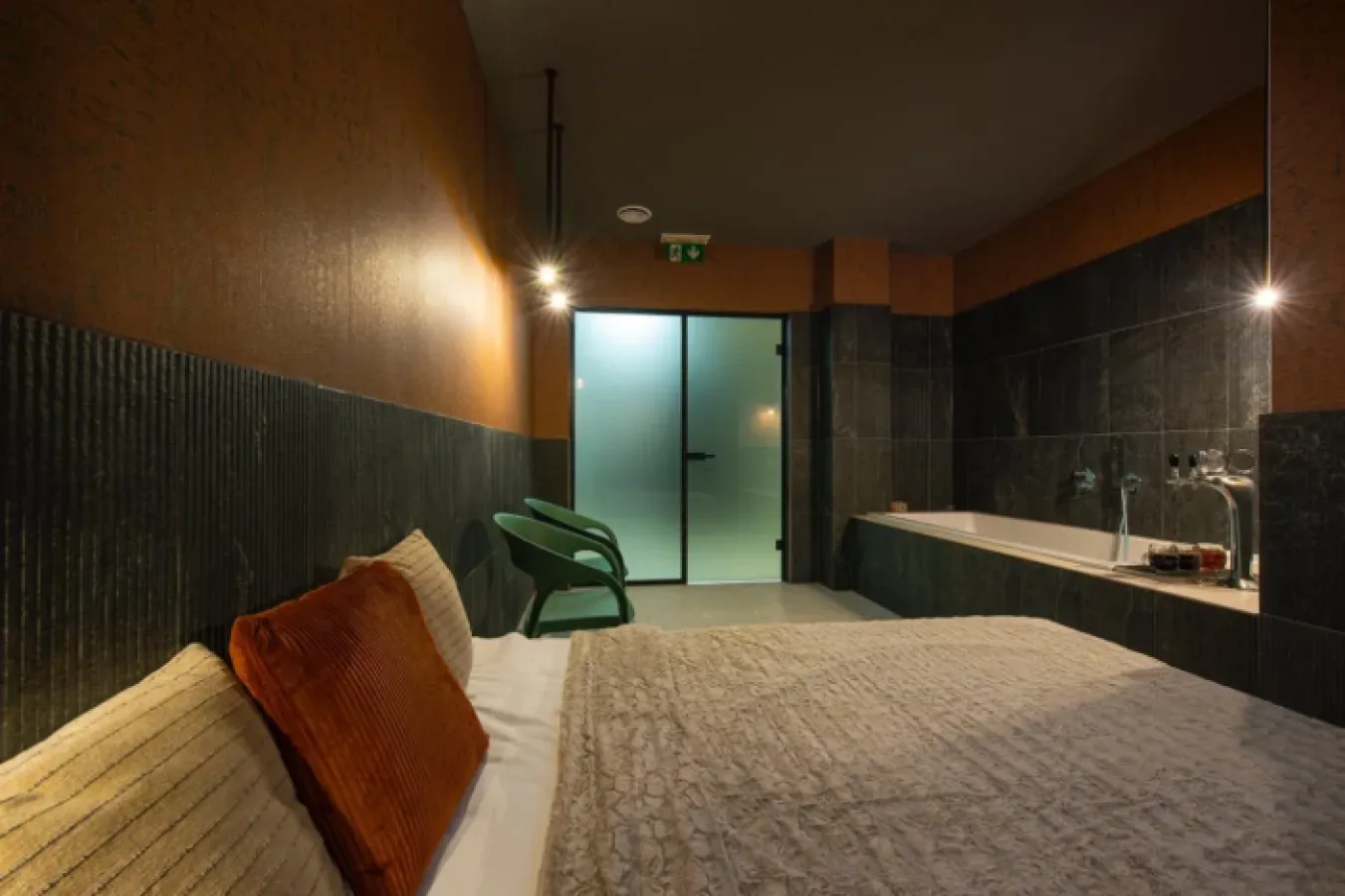 A dimly lit bath room with a bed, a tub in dark tiling and a frosted glass door
