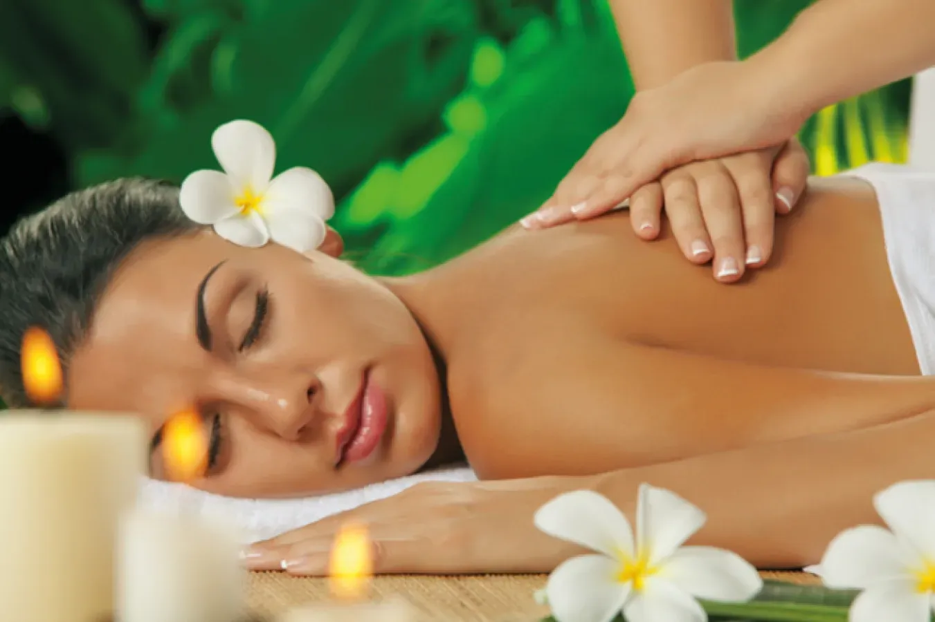 A woman with a flower in her hair having a back massage, with lit candles and frangipani flowers in front