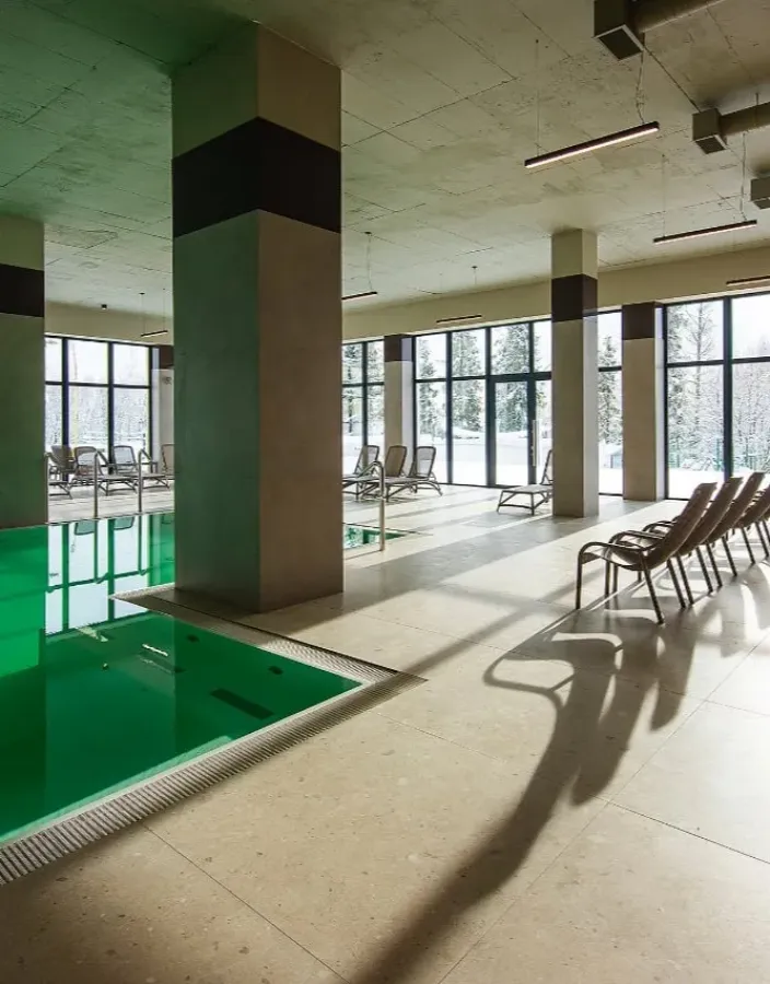 An indoor swimming pool with green water, a row of loungers and a glass wall onto the forest