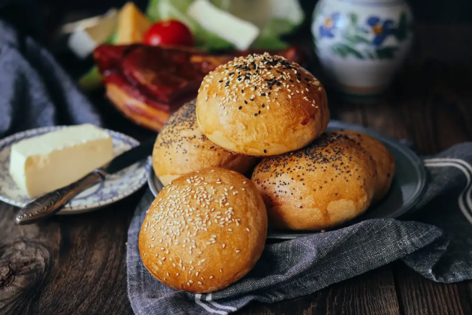 Fresh sesame and poppy seed rolls on a tray, with butter and smoked meat beside them